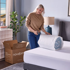 A young woman cutting the plastic on the still compressed topper on top of her mattress. An open delivery box sits on the floor behind her.