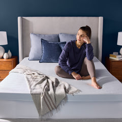 The young woman comfortably sits up in bed, softly smiling with the topper supporting her body. The bedroom is sunny with navy blue walls and various blue accents.