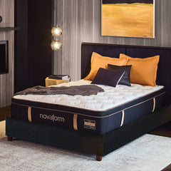 Front angled view of the Novaform Legacy Premier Support Hybrid Mattress in a bedroom against a wood paneled wall. There are gold pillows on the bed and a fireplace on the opposite wall. 