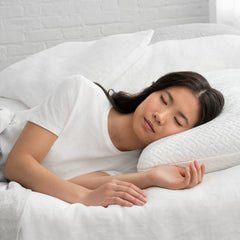 Young woman lying on her side asleep in bed with her head resting on the Lasting Cool Foam Pillow her left hand touching the cool cover