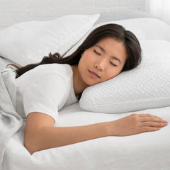 Young woman lying on her stomach asleep in bed with her head resting on the Lasting Cool Pillow and her right arm resting on the mattress