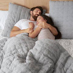 Young man lying on his back cuddling a young woman lying on her side sleeping together in pajamas with a gray comforter pulled up to their waists over the Serafina Pearl mattress cover