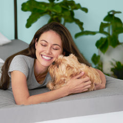 Young woman lying in bed on her stomach smiling and cuddling a small tan dog kissing her face