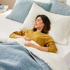 Young woman wearing pajamas lying on her back with her head propped up against a pillow and her hands on the comforter