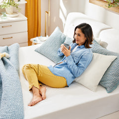 Young woman wearing daytime clothing sitting up in bed against a bunch of pillows drinking a cup of coffee