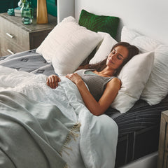 Young woman in pajamas asleep on her back with her head and neck propped up on white pillows with the comforter pulled up below her chest and the charcoal Noir Mattress cover revealed underneath