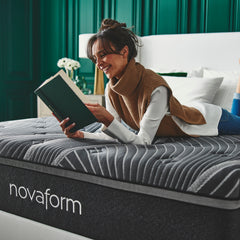 Young woman wearing day clothes lying on her stomach on top of the Noir Mattress while reading a book and smiling