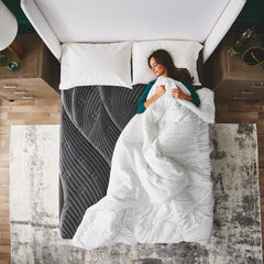 Overhead view of a woman in pajamas lying asleep on her back in bed with a white comforter pulled up to her neck on the quilted fiberfill mattress cover